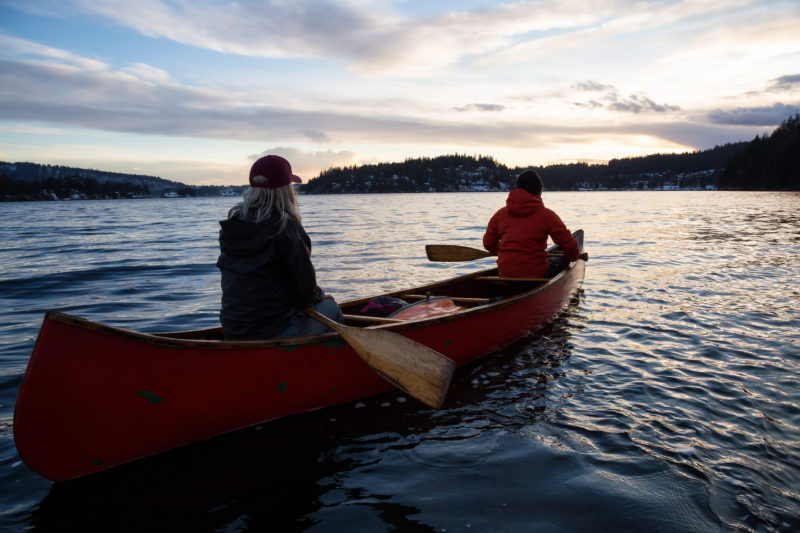 Couple friends on a wooden canoe are paddling in an inlet surrounded by Canadian mountains during a vibrant sunset. Taken in Indian Arm, near Deep Cove, North Vancouver, British Columbia, Canada.